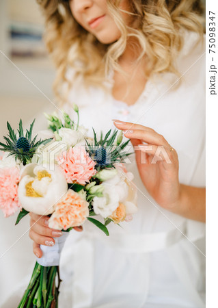 The bride holds in her hand and gently touches a bouquet with peonies, roses, lisianthus and eryngiums The bride holds in her hand and gently touches a bouquet with peonies, roses, lisianthus and eryngiums 75098347