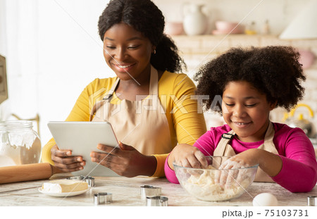 Cheerful Black Mom And Daughter Using Digital Tablet In Kitchen, Checking Recipe 75103731