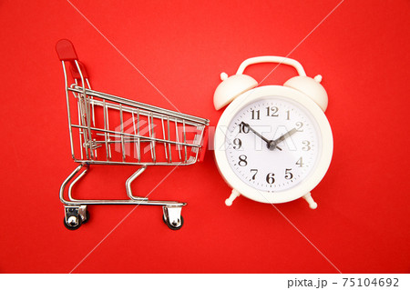 Mini shopping cart with white alarm clock on red background. Conceptual image of sale, seasonal discounts in shopping stores, discount time, Time saving concept Mini shopping cart with white alarm clock on red background. Conceptual image of sale, seasonal discounts in shopping stores, discount time, Time saving concept 75104692