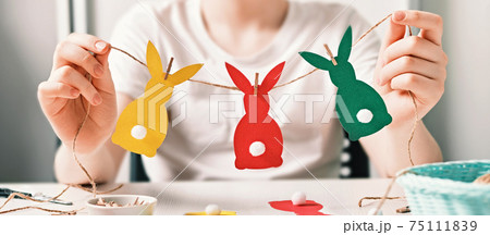 Banner with DIY Easter home decor. Happy boy make garland of colored paper, twine and pom-poms. Selective focus Banner with DIY Easter home decor. Happy boy make garland of colored paper, twine and pom-poms. Selective focus 75111839