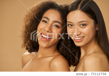Portrait of two joyful attractive mixed race young women with perfect smile posing for camera isolated over beige background 75112756