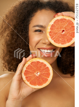 Portrait of mixed race young woman looking happy while posing with grapefruit cut in half over beige background 75112759