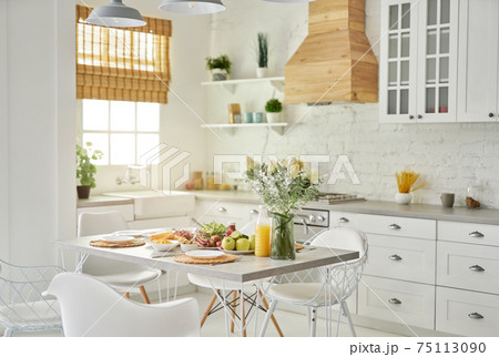 Cozy kitchen. Modern bright white kitchen interior with wooden and white details. Vase with flowers and Latin style breakfast on the table 75113090