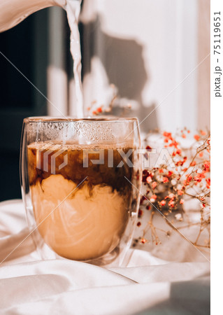 Milk pouring into the glass cup with black coffee. Morning aesthetics vibes. Breakfast. Pink gypsophila flowers 75119651