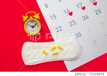 Menstruation calendar with pads, alarm clock, hormonal contraceptive pills on red background. Female's menstrual cycle concept. Pain relievers for menstrual pain 75120028