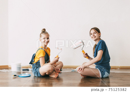 A girl with her sister in bright blue and yellow clothes helps to paint the walls in her room white A girl with her sister in bright blue and yellow clothes helps to paint the walls in her room white 75125382