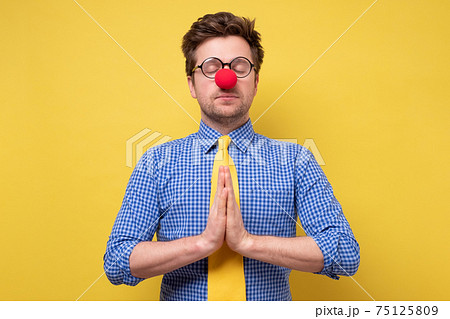 Funny young man with red clown nose pressings palms together, trying to calm down. 75125809