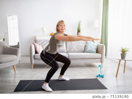 Home workout concept. Beautiful mature lady in sportswear doing squats on yoga mat indoors, free space Home workout concept. Beautiful mature lady in sportswear doing squats on yoga mat indoors, free space 75139190