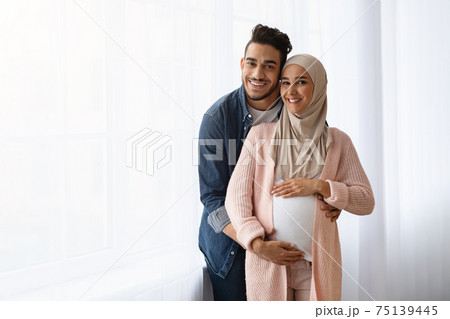 Pregnant Muslim Woman And Her Husband Embracing Near Window At Home 75139445