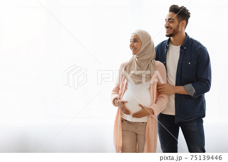 Pregnant Religious Muslim Couple Standing Near Window At Home, Looking Aside Pregnant Religious Muslim Couple Standing Near Window At Home, Looking Aside 75139446