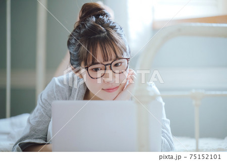寝室のベッドに寝そべるパソコンを操作するメガネをかけたお団子ヘアーの若い女性 75152101