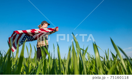 Wind swayes U.S. flag in the hands of a woman standing on a green wheat field 75160118