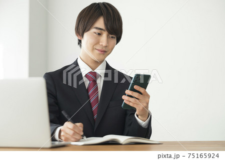 スマートフォンの画面を見ながらメモをとるスーツ姿の若い男性 75165924
