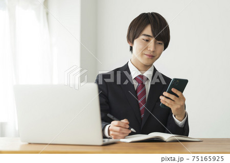 スマートフォンの画面を見ながらメモをとるスーツ姿の若い男性 スマートフォンの画面を見ながらメモをとるスーツ姿の若い男性 75165925