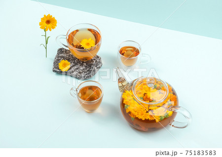 Cups of herbal tea, transparent teapot with calendula flowers and stones on blue background. Calendula Tea Benefits Your Health concept. Copy space Cups of herbal tea, transparent teapot with calendula flowers and stones on blue background. Calendula Tea Benefits Your Health concept. Copy space 75183583