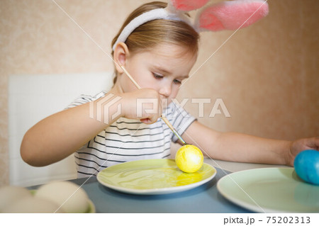 little cute blue-eyed girl in bunny ears paints Easter eggs 75202313