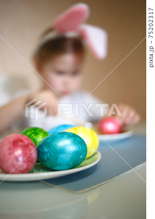 little cute blue-eyed girl in bunny ears paints Easter eggs 75202317