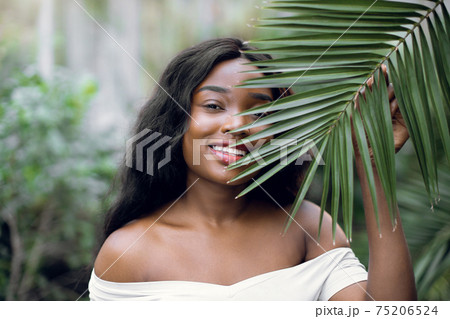 Fashion tropical portrait. Young attractive dark skinned woman model in white shirt, holding in hands green palm leaf while posing in beautiful greenhouse wth tropical plants Fashion tropical portrait. Young attractive dark skinned woman model in white shirt, holding in hands green palm leaf while posing in beautiful greenhouse wth tropical plants 75206524