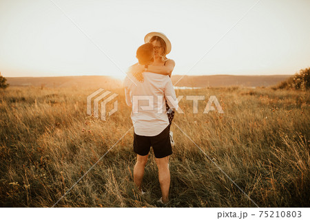 A man with a woman in a hat hug and kiss in the tall grass in the meadow 75210803