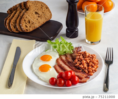 Hearty breakfast with scrambled eggs, bacon, beans, cherry tomatoes, homemade bread, orange juice on a concrete countertop. English breakfast concept. Hearty breakfast with scrambled eggs, bacon, beans, cherry tomatoes, homemade bread, orange juice on a concrete countertop. English breakfast concept. 75211000
