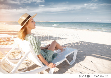 Young attractive caucasian woman in a green dress and a straw sits resting on a lounger under a wooden umbrella on a sandy beach of the seashore or ocean. 75236236