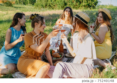Friends having picnic in the countryside. Group of young women sitting on blanket in park near trees, at sunset on spring summer day. Five girlfriends eating and drinking red wine on outdoor party Friends having picnic in the countryside. Group of young women sitting on blanket in park near trees, at sunset on spring summer day. Five girlfriends eating and drinking red wine on outdoor party 75240148