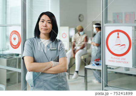 Young cross-armed female clinician in uniform standing against colleague and patient Young cross-armed female clinician in uniform standing against colleague and patient 75241899