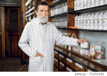 People, healthcare, pharmacy concept. Indoor portrait of pleasant professional mature bearded man pharmacist, wearing white coat, posing in beautiful old pharmacy interior 75243162