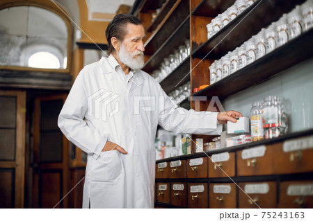 Portrait of handsome bearded senior man pharmacist, working at vintage old pharmacy, taking box with pills from wooden shelf Portrait of handsome bearded senior man pharmacist, working at vintage old pharmacy, taking box with pills from wooden shelf 75243163