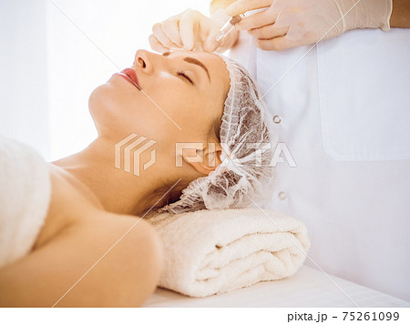 Beautiful woman receiving beauty injections with closed eyes in sunny medical center. Beautician doctor hands doing beauty procedure to female face with syringe. Cosmetic medicine and surgery concept Beautiful woman receiving beauty injections with closed eyes in sunny medical center. Beautician doctor hands doing beauty procedure to female face with syringe. Cosmetic medicine and surgery concept 75261099
