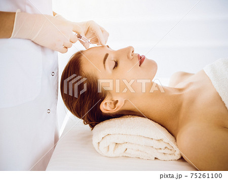 Beautiful woman receiving beauty injections with closed eyes in sunny medical center. Beautician doctor hands doing beauty procedure to female face with syringe. Cosmetic medicine and surgery concept 75261100