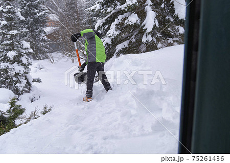 Snow removal on the roof house. Boy in green jacket with shovel throws snow off the roof. Snow removal on the roof house. Boy in green jacket with shovel throws snow off the roof. 75261436