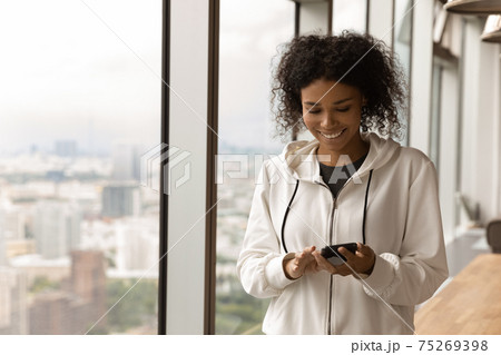 Smiling African American woman using phone, standing near panoramic window 75269398
