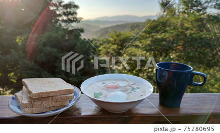 rice porridge with pork and boiled egg with coriander leaf on white bowl serving with black mug of home made coffee and grilled Whole wheat bread in the middle of forest mountain in Summer season, adv 75280695