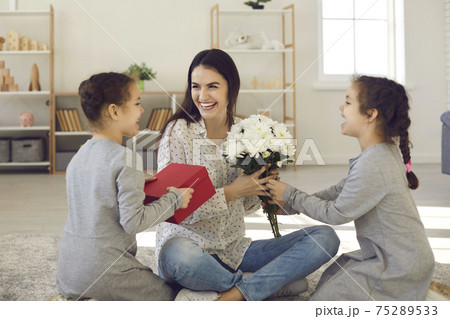 Happy smiling woman mother sitting on floor between her daughters and getting flowers and presents Happy smiling woman mother sitting on floor between her daughters and getting flowers and presents 75289533