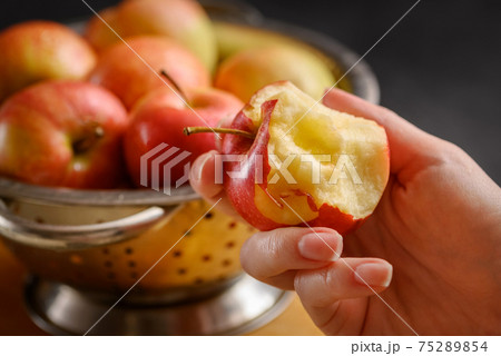 Bitten apple in human hand on metallic bowl full of red ripe apples background. Healthy eating. Apple pie ingridients. Cooking at home Bitten apple in human hand on metallic bowl full of red ripe apples background. Healthy eating. Apple pie ingridients. Cooking at home 75289854