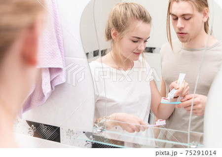 Young married couple of teenagers at the mirror in the bathroom getting ready to brush their teeth by squeezing the paste on each other's toothbrushes 75291014