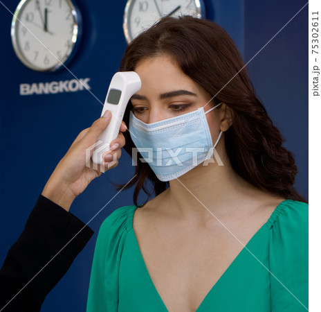 A Brazilian tourist in green clothes wear a mask while measuring the temperature with a fever monitor diagnostic tool. The back wall has a clock to tell the time in the capitals of different countries 75302611