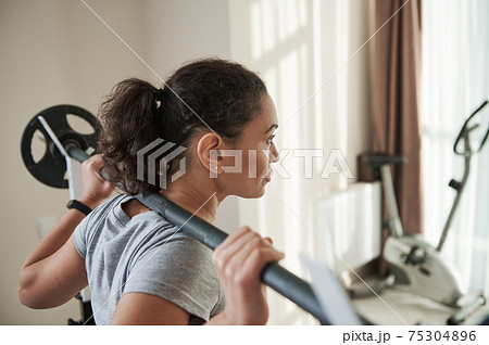 Rear view of African young muscular woman squatting with barbell at home gym. Rear view of African young muscular woman squatting with barbell at home gym. 75304896