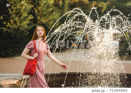 Young beautiful red-haired long-haired woman in a summer pink dress walks through the streets of a summer city. 75304905