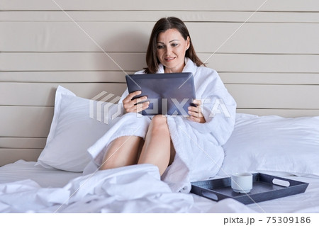 Mature female in white bathrobe with cup of coffee looking at webcam of digital tablet 75309186