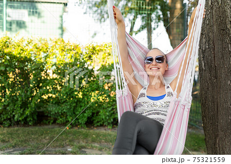 Young adult happy short haired shaved bald attractive woman chilling enjoy relaxing sitting in fabric chair hammock at yard in garden near house. Peaceful and idyllic outdoor rest lifestyle Young adult happy short haired shaved bald attractive woman chilling enjoy relaxing sitting in fabric chair hammock at yard in garden near house. Peaceful and idyllic outdoor rest lifestyle 75321559
