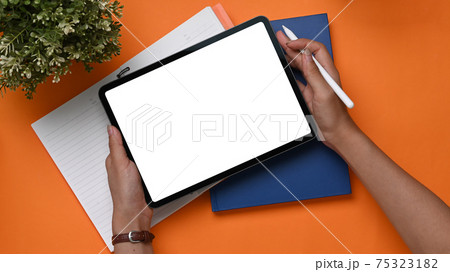 Overhead shot of young woman hands holding digital tablet and stylus pen on orange background. Overhead shot of young woman hands holding digital tablet and stylus pen on orange background. 75323182