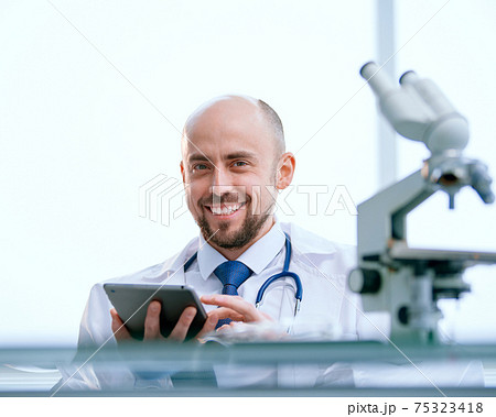 smiling researcher with a digital tablet sitting in front of a microscope smiling researcher with a digital tablet sitting in front of a microscope 75323418