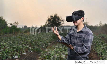 Young asian man smart farmer standing in green field and wearing visual reality glasses technology. 75324590