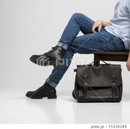 Man in a blue jeans and black boots sits on a chair with a brown men's shoulder leather bag for a documents and laptop on a white floor. Mens leather satchel, messenger bags, handmade briefcase. Man in a blue jeans and black boots sits on a chair with a brown men's shoulder leather bag for a documents and laptop on a white floor. Mens leather satchel, messenger bags, handmade briefcase. 75326289