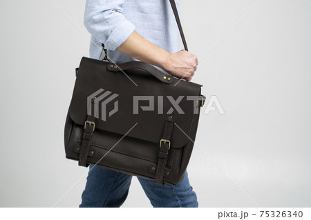 Brown men's shoulder leather bag for a documents and laptop on the shoulders of a man in a blue shirt and jeans with a white background. Satchel, mens leather handmade briefcase. Brown men's shoulder leather bag for a documents and laptop on the shoulders of a man in a blue shirt and jeans with a white background. Satchel, mens leather handmade briefcase. 75326340