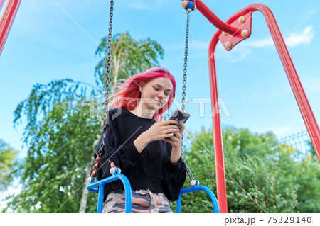 Fashionable happy teenage girl with dyed hair with smartphone riding swing 75329140