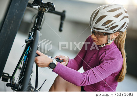 Concentrated female cyclist wearing protective helmet and glasses looking focused while using pump for inflating the tire of her bicycle, kneeling outdoors on a daytime Concentrated female cyclist wearing protective helmet and glasses looking focused while using pump for inflating the tire of her bicycle, kneeling outdoors on a daytime 75331092