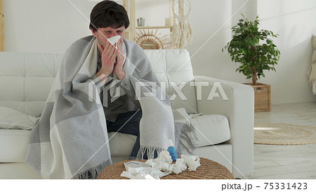 Man wrapped in a blanket feels sick with a cold and fever at home, sick with the flu, sitting on the couch Man wrapped in a blanket feels sick with a cold and fever at home, sick with the flu, sitting on the couch 75331423